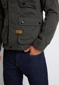 Oliwkowa kurtka G-Star z wieloma kieszeniami z przodu, zapięciem na guziki i wytłoczonym logo. Połączona z ciemnoniebieskimi dżinsami.