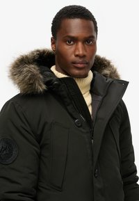 Zwarte parka jas met een afneembare capuchon van faux bont, rits- en knoopsluiting, en een gebreide crèmekleurige hoge collar. Inclusief logo patch op de mouw.