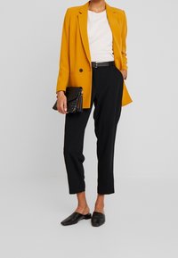 Kobieta w musztardowej marynarce, białej koszulce, czarnych spodniach o długości 3/4, czarnym pasku, czarnych mokasynach, trzymająca czarną teksturowaną kopertówkę.