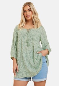 Lös passform på grön blus med blommönster, rund halslinje, knytdetalj och 3/4-långa ärmar. Kombinerad med blå denimshorts.