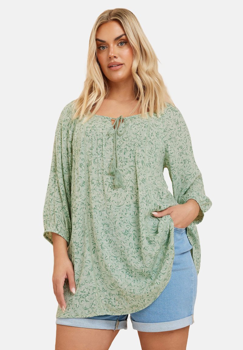 Lös passform på grön blus med blommönster, rund halslinje, knytdetalj och 3/4-långa ärmar. Kombinerad med blå denimshorts.