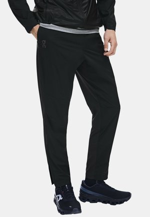 Homme portant un pantalon de sport noir, des chaussures de course noires, un t-shirt gris et une veste noire avec une main dans la poche.