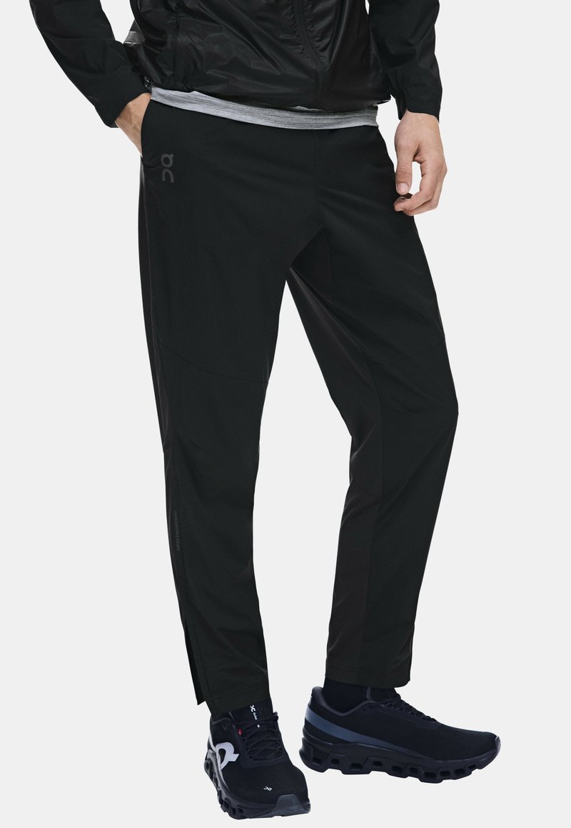 Homme portant un pantalon de sport noir, des chaussures de course noires, un t-shirt gris et une veste noire avec une main dans la poche.
