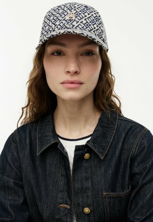 Giovane donna con capelli castani ondulati, che indossa un berretto decorato con un piccolo emblema dorato e una giacca di denim scuro con bottoni dorati.