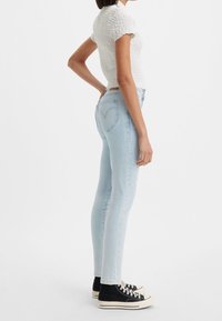 Jeans de mezclilla skinny en azul claro con cintura alta y bolsillos traseros, combinados con una blusa blanca texturizada de manga corta y zapatillas negras.