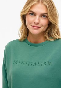 Femme blonde aux yeux bleus portant un sweat-shirt vert brodé du mot "MINIMALISME" sur la poitrine, souriant légèrement.