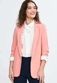 Brzoskwiniowa marynarka o strukturze i podwiniętych rękawach, noszona na białej bluzce z kokardą. Ciemne spodnie dopełniają strój.