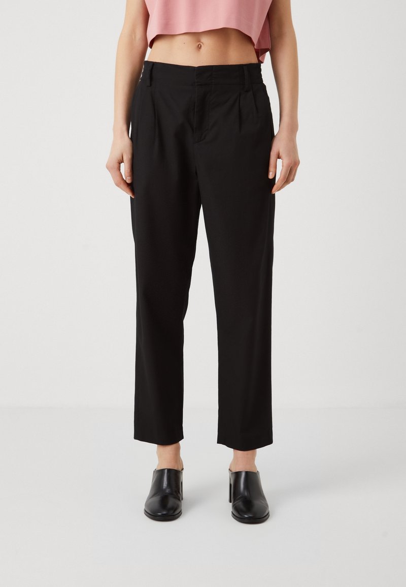 Femme portant un pantalon noir taille haute sur mesure, des chaussures à talons noires sans lacets et un top rose court, posant devant un fond clair uni.