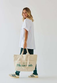 Beżowa torba płócienna z graficznym napisem "WITAJ W KLUBIE PILATESU" w ciemnozielonym kolorze. Modelka nosi białą, oversize'ową koszulę i zielone legginsy.
