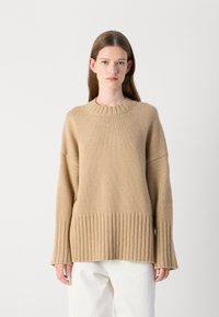 Beige gebreid sweater met een geribbelde zoom en manchetten, met een ronde halslijn en een losse pasvorm. Het materiaal lijkt zacht en gestructureerd.
