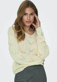 Maglione a maglia giallo chiaro con vestibilità ampia, caratterizzato da un collo a V e un motivo di punto aperto. Indossato sopra pantaloni grigi scuri.