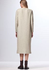 Strickkleid mit langen Ärmeln in hellbeige, mit gerader Schnittführung und seitlichen Schlitzen; getragen mit dunkelbraunen Stiefeln, die einen Kontrast setzen.