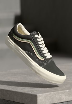 Black and white low-top sneaker with white laces and green and beige stripe detailing, suspended above a concrete surface.