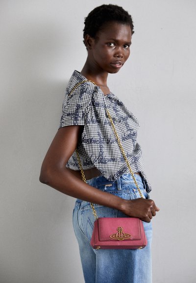 Rød læder crossover-taske med en gylden kædestrop, der har et karakteristisk design af gyldent hardware. Modellen bærer en mønstret crop top og denimjeans.