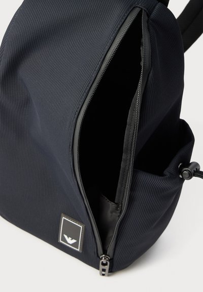 Μαύρο υφασμάτινο σακίδιο με υφή, με ανοιχτή τσέπη με φερμουάρ και ετικέτα λογότυπου στο μπροστινό μέρος, που δείχνει μια πλαϊνή τσέπη με ρυθμιζόμενο κορδόνι.