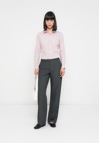 Vrouw staat opgesteld met een roze gestreepte blouse, grijze broek, zwarte schoenen en houdt een witte handtas vast tegen een eenvoudige witte achtergrond.