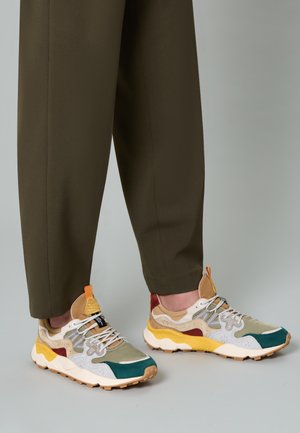 Person wearing olive pants and multicolored athletic sneakers with yellow, green, beige, and white accents standing on a gray floor.