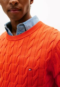 Maglione arancione a lavorazione a trecce con scollo a coste, indossato sopra una camicia di jeans blu chiaro. Piccola etichetta con logo visibile sul maglione.