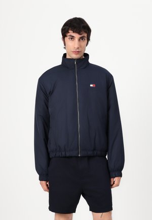 Marineblauwe ritsjack met hoge kraag, elastische tailleband en manchetten, met een klein meerkleurig logo op de borst.