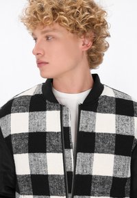 Czarna i biała bomberka w kratę z zamkiem z przodu, dzianinowym kołnierzem i wzorzystą fakturą na przodzie, kontrastującą z czarnymi rękawami.