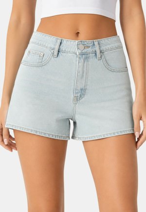 Donna che indossa shorts di jeans azzurro chiaro e un top corto bianco, pancia scoperta, in piedi con le braccia rilassate lungo i fianchi.