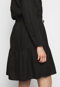 Femme portant une robe noire à manches longues avec une taille froncée et une jupe à volants, montrant le côté et les détails du bras jusqu'à mi-hauteur.