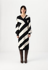 Robe tricotée avec des rayures diagonales noires et crème, décolleté en V, manches surdimensionnées et longueur midi. Des bottes à motifs marron complètent le look.