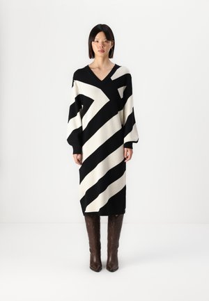 Rochie tricotată cu dungi diagonale negre și crem, decolteu în V, mâneci supradimensionate și lungime până la jumătatea gambei. Botine din piele cu model maro completează look-ul.