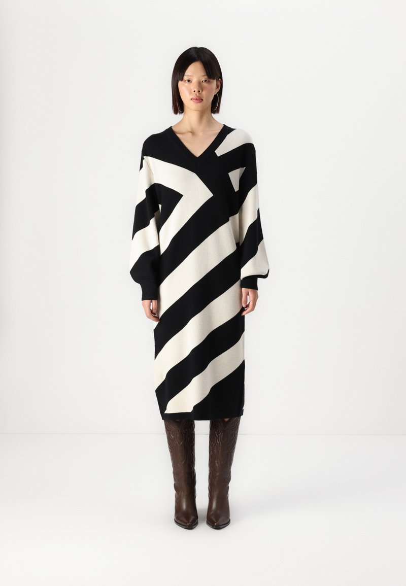 Robe tricotée avec des rayures diagonales noires et crème, décolleté en V, manches surdimensionnées et longueur midi. Des bottes à motifs marron complètent le look.
