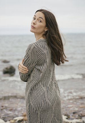 Femme aux longs cheveux bruns portant un pull gris à motifs, debout au bord d'un rivage rocheux, regardant par-dessus son épaule en direction de la caméra.