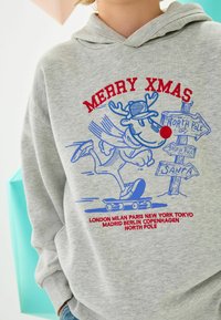 Grå hættejakke med et blåt og rødt grafisk motiv af et rensdyr på et skateboard. Teksten siger "GLÆDELIG JUL" og bynavne med vejskilt.