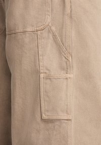 Beige cargo shorts gjorda av slitstark bomullsmaterial, med sidofickor och en texturerad tyg med synliga sömmar.