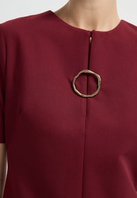 Robe bordeaux à manches courtes, présentant un fermoir doré texturé en forme ronde au niveau du cou et une couture droite sur le devant.