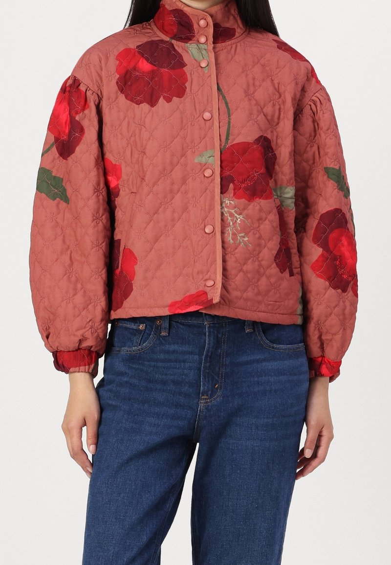 Kvinde iført en kviltet støvet rosa jakke med store røde blomsterprints og puffy ærmer, parret med blå denimjeans.