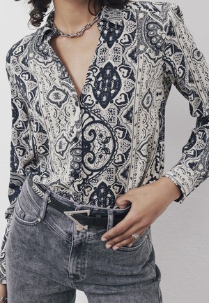 Femme portant une chemise boutonnée à motifs noir et blanc, un jean taille haute gris, une ceinture noire avec une boucle triangulaire argentée, et un collier chaîne argenté.