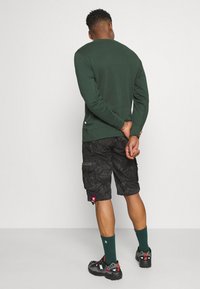 Felpa verde scuro con collo rotondo, abbinata a pantaloni cargo neri mimetici. Sneakers nere e calze verdi completano l'outfit.