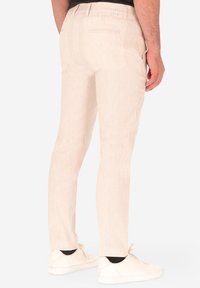 Uomo che indossa pantaloni a righe beige chiaro e sneakers bianche, visto da dietro contro uno sfondo bianco uniforme.