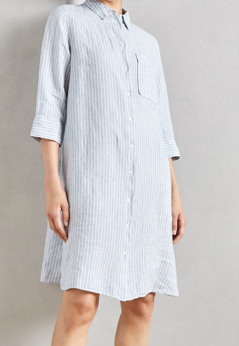 Femme portant une robe chemisier à rayures verticales bleu clair et blanc, avec des manches trois-quarts et une poche poitrine, debout devant un fond uni.