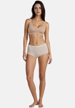 Kvinde stående barfodet iført beige sømløs bh og trusser med høj talje mod en ensfarvet hvid baggrund.