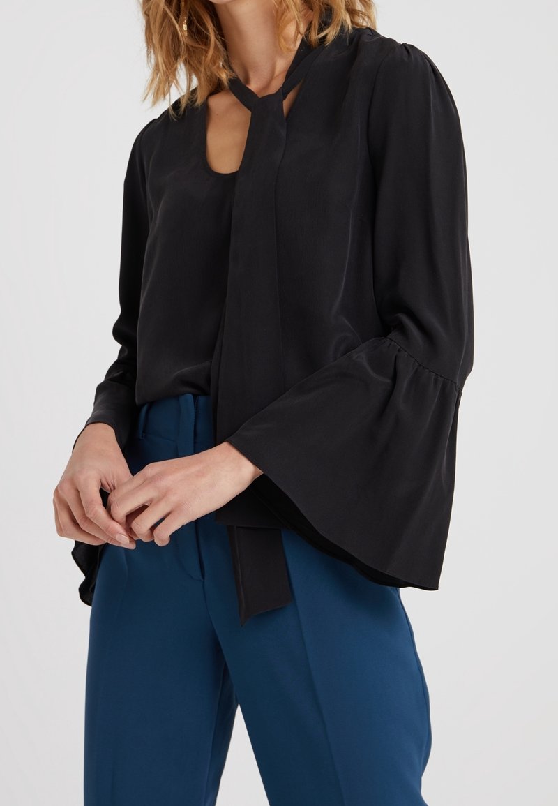 Femme portant un chemisier noir à manches longues avec des manches évasées et un nœud au col, associé à un pantalon bleu taille haute.