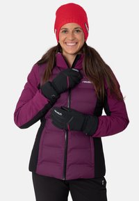 Mujer sonriente que lleva un gorro de lana rojo, una chaqueta de invierno púrpura y negra, y guantes negros, posando con las manos cerca del zipper de la chaqueta.