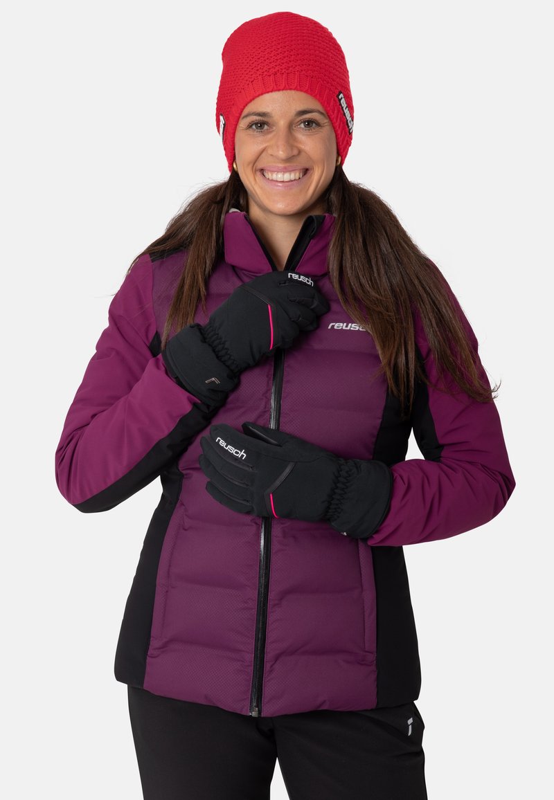 Lächelnde Frau trägt eine rote Strickmütze, eine lila-schwarze Winterjacke und schwarze Handschuhe, posiert mit den Händen nahe dem Reißverschluss der Jacke.
