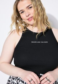 Top in maglia nera con costine presenta la scritta ricamata in bianco "DOVE SEI ORA". Il modello indossa intricati anelli mentre sfoggia una gonna fantasia.