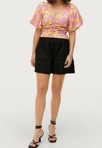 Blumiges Crop-Top mit kurzen Puffärmeln in Pink und Gelb, kombiniert mit schwarzen Leinen-Shorts. Schwarze Riemchen-Highheels runden das Outfit ab.