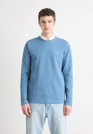 Chemise à manches longues bleu clair avec un col rond et une texture côtelée, arborant un petit logo "BOSS" sur la poitrine. Associée à un jean en denim clair.