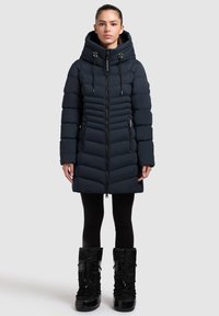 Temno modra puhasta jakna z kapuco, s cik-cak črtnim vzorcem, zadrgo spredaj in z dodatki z zavezovanjem; nošena čez črne legice in škornje.