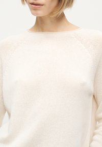 Femme portant un pull en tricot de couleur crème avec un col rond, montrant le haut du torse et une partie du visage.