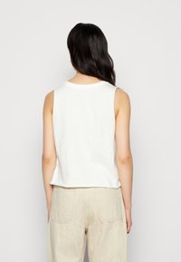 Top bianco senza maniche con scollatura rotonda, realizzato in tessuto morbido, caratterizzato da un taglio corto e un orlo leggermente curvo. Abbinato a pantaloni beige chiaro.