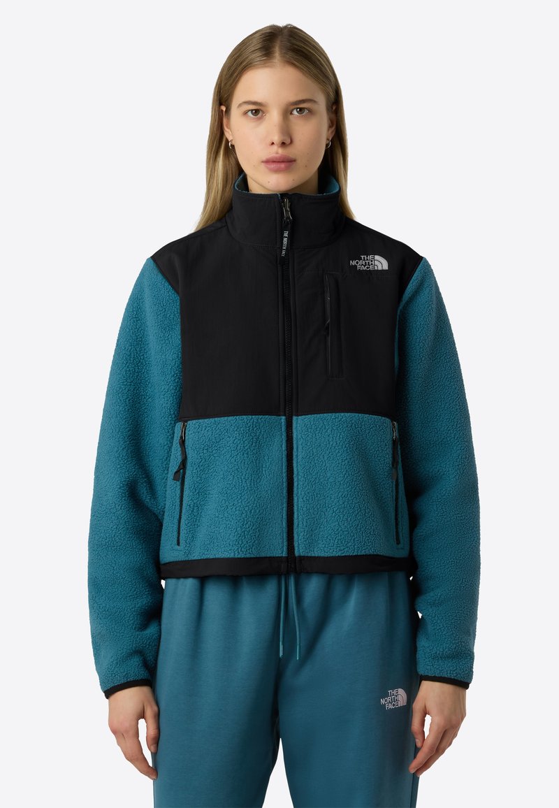 Parte superiore di una giacca in pile color teal con parte superiore in nylon nero, dotata di una tasca sul petto con zip e due tasche laterali con zip.