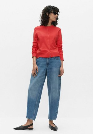 Vrouw met krullend haar die een zwarte zonnebril draagt, een rood vest met knopen, losse blauwe spijkerbroek en zwarte slingback ballerina's, zijwaarts staand.